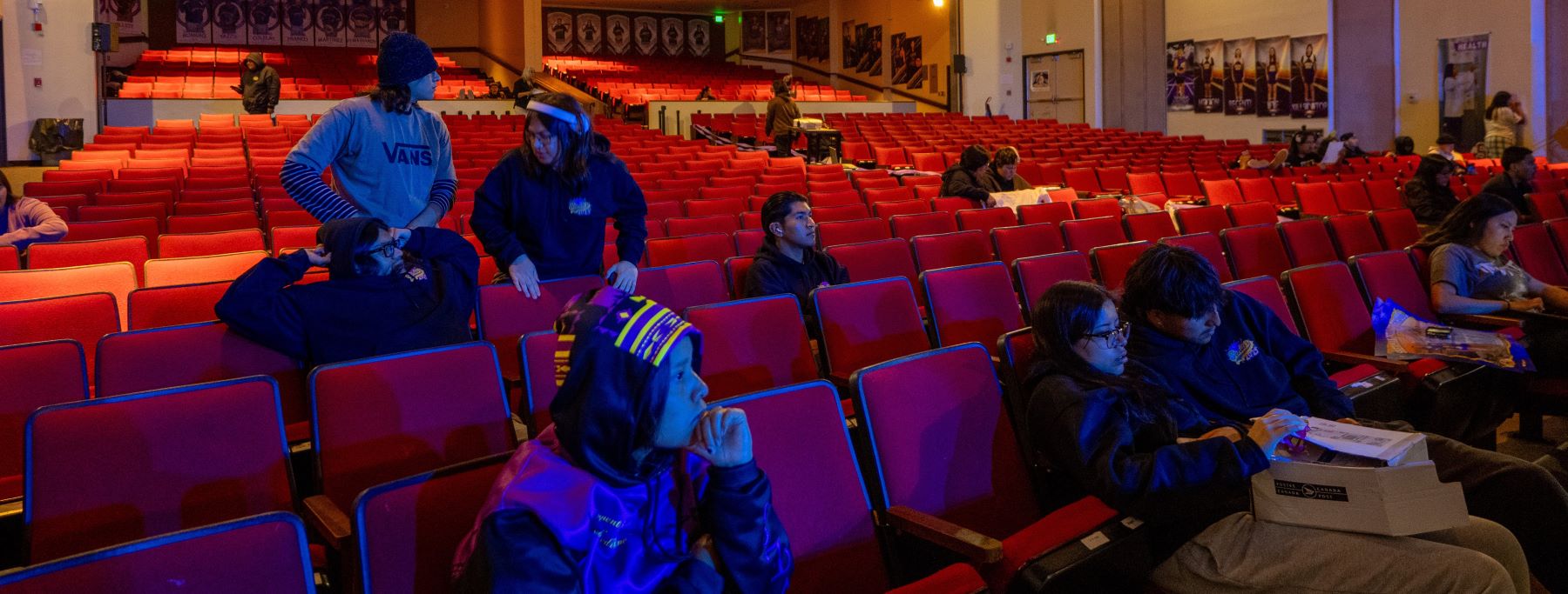Students sitting in red seats inside an auditorium, engaged in an event.
