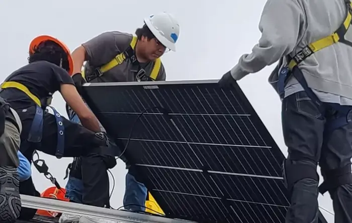 Three workers wearing safety harnesses and helmets install a large solar panel on a rooftop. One person holds the panel steady while the others assist in positioning it.