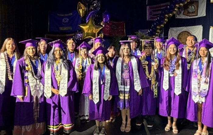 A group of graduating seniors from Sherman Indian High School pose together on stage in purple caps and gowns.