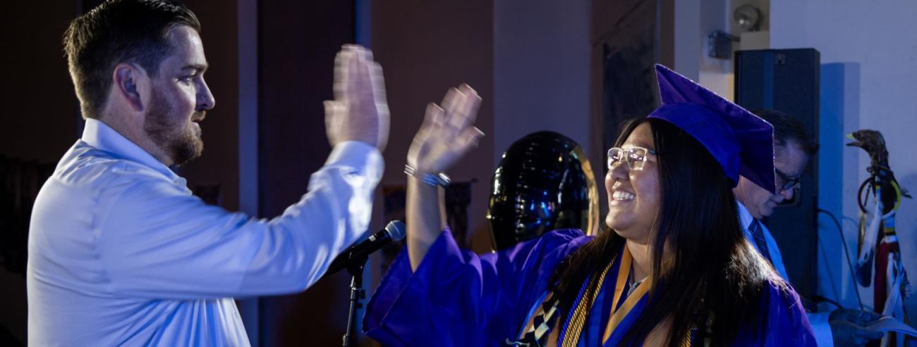 A school staff member and student in graduation gowns high-fiving each other, celebrating their achievement.