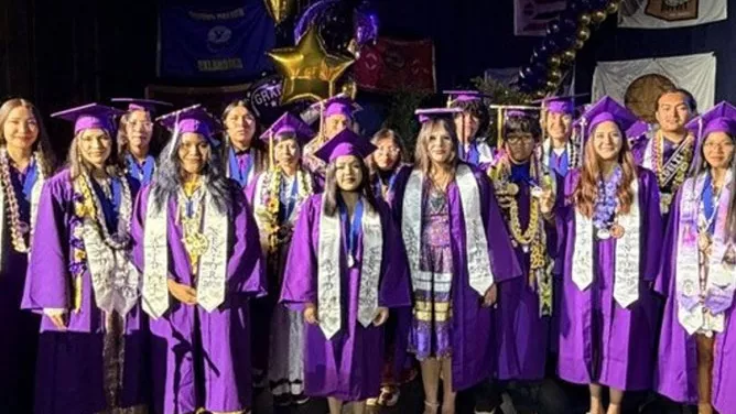 A group of graduating seniors from Sherman Indian High School pose together on stage in purple caps and gowns.